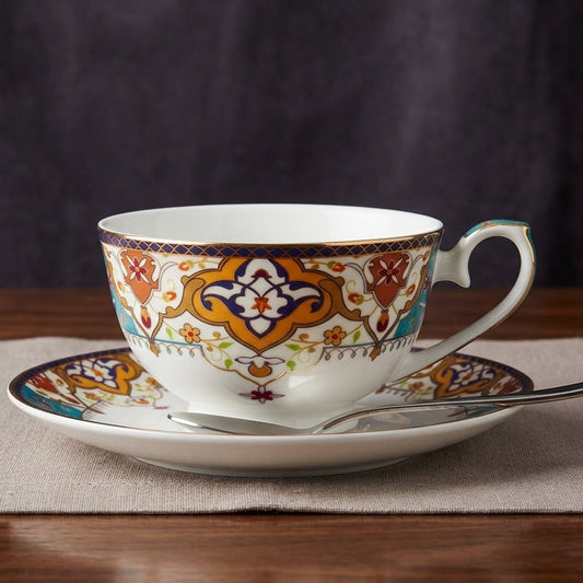 A white porcelain teacup and saucer set with intricate orange, blue, and gold geometric patterns, displayed on a beige placemat over a wooden table.