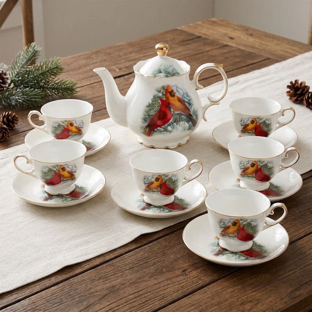 A white porcelain teapot and matching teacups with saucers, decorated with red cardinal birds and green pine branches, arranged on a wooden table with a white runner and holiday greenery.
