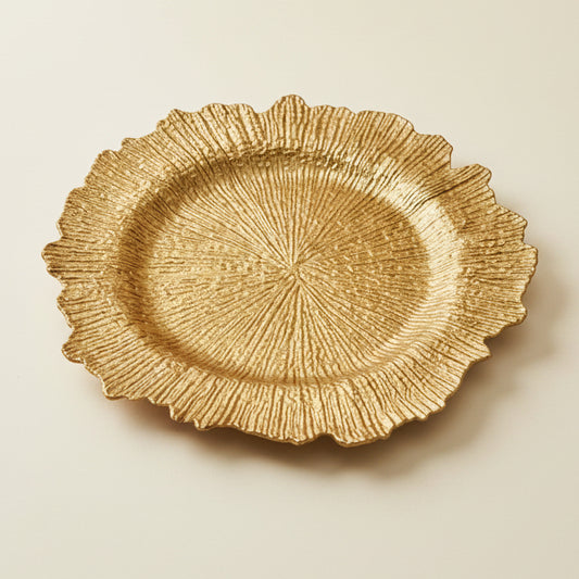A collage showing a gold-toned decorative plate with a textured, radiating leaf-like scalloped edge. The plate is shown from multiple angles: on a dark wood surface, on a white marble surface, held by a hand in a warehouse setting, and in a close-up detail view highlighting the metallic texture.