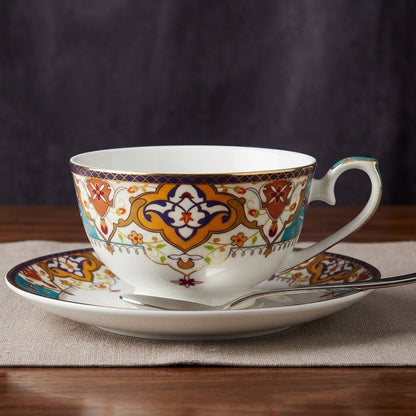 A white porcelain teacup and saucer set with intricate orange, blue, and gold geometric patterns, displayed on a beige placemat over a wooden table.