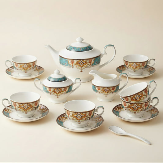 A ceramic tea and coffee cup set arranged on a tabletop, including a teapot, sugar bowl, creamer, teacups, saucers, and a small copper-colored tea strainer. The white porcelain is decorated with ornate gold, turquoise, and red geometric patterns, set against a neutral indoor background.