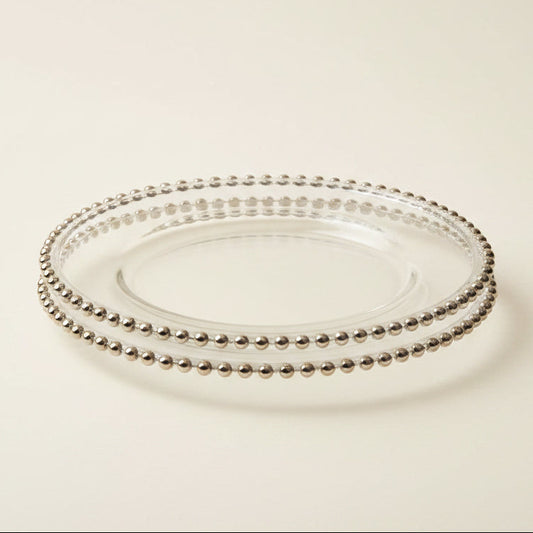 Three-panel collage showing a clear glass charger plate with a silver beaded rim, first close-up on white fabric, then stacked on a dark wood table, and finally styled on a table setting with white plates, silverware, flowers, and candles.
